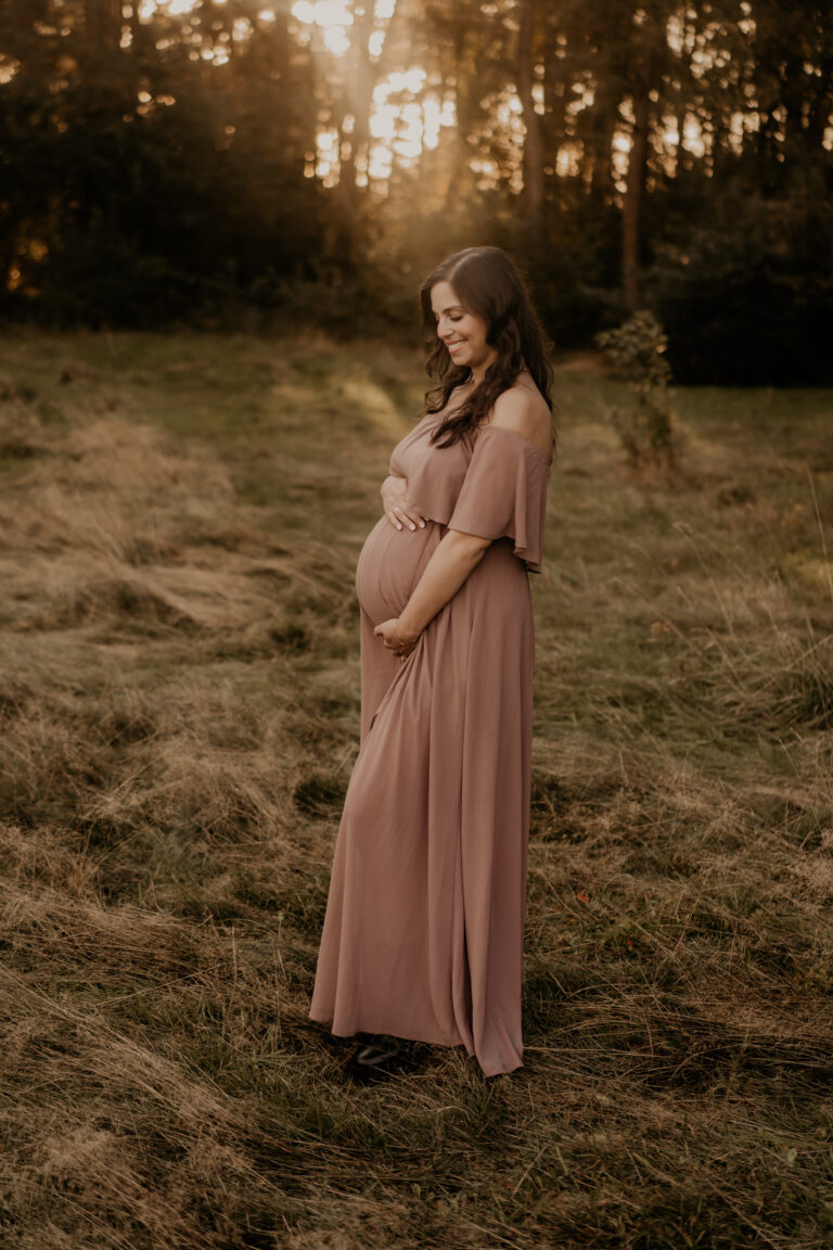 Schwangerschaftsfoto im Abendlicht – Eine werdende Mutter in einem fließenden, altrosafarbenen Kleid hält ihren Babybauch sanft und lächelt