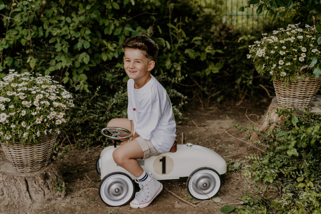Lächelnder Junge sitzt auf einem weißen Retro-Tretauto im Garten, umgeben von Blumen – Kindergartenfoto von Kita-Fotografin in Lengerich