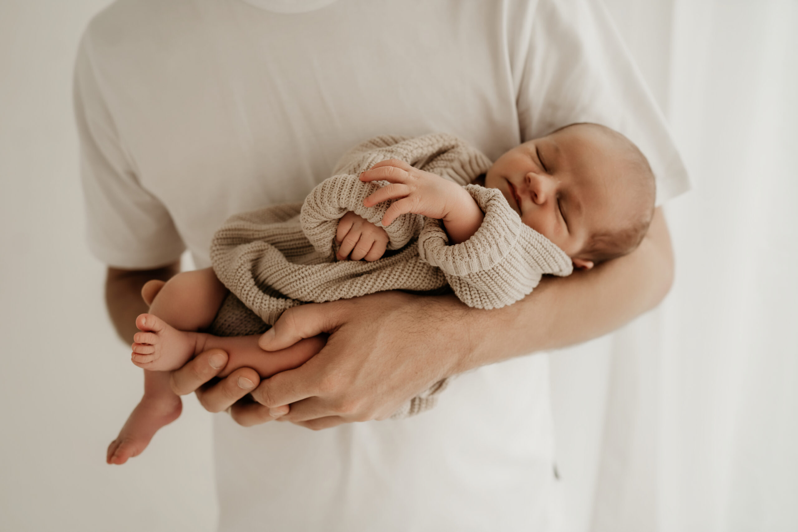 Papa hält sein Baby sicher und liebevoll. Minimalistische und emotionale Neugeborenenfotografie mit Fokus auf echte Verbindung.