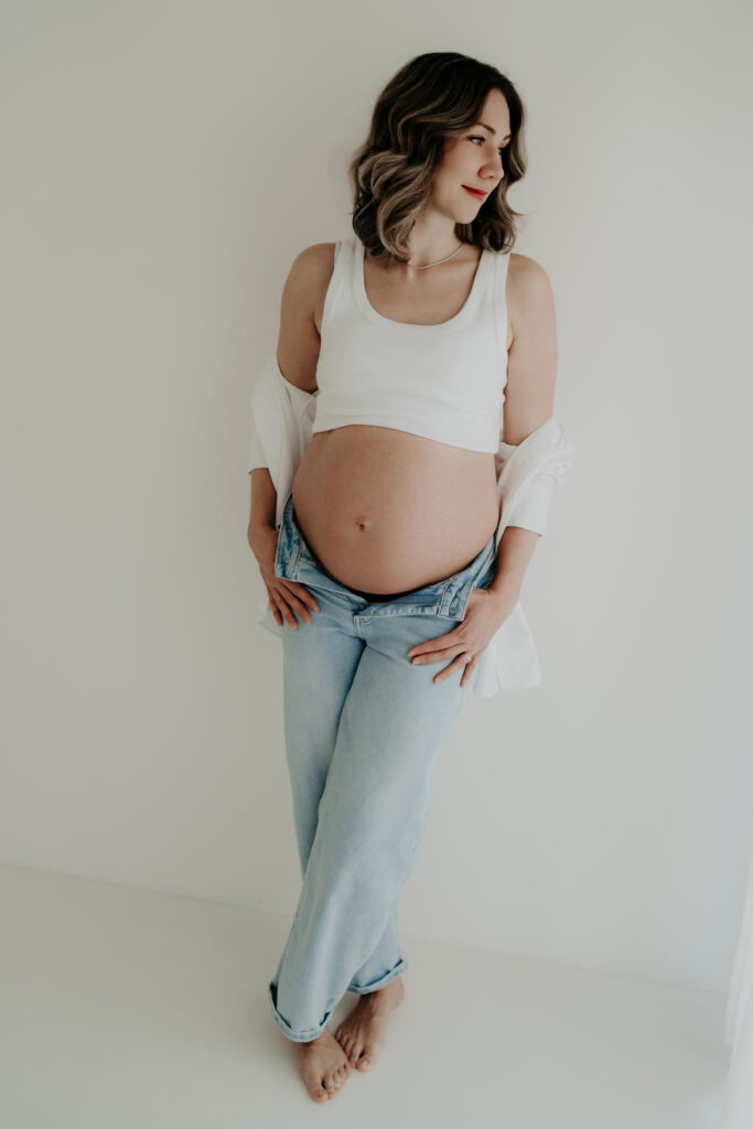 Schwangere Frau steht im hellen Studio mit Jeans und schaut zur Seite