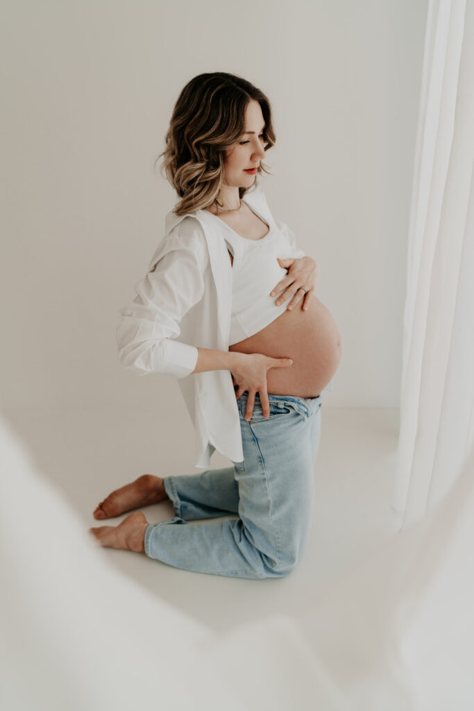 Schwangere Frau kniet im Profil im hellen Fotostudio und hält ihren Bauch