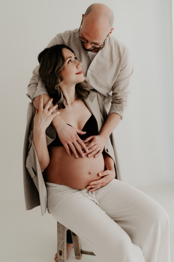 Schwangere Frau sitzt auf Hocker und schaut ihren Partner liebevoll an im Fotostudio