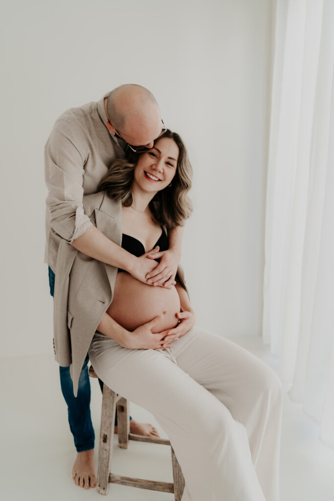 Paar beim Babybauchshooting im hellen Studio, Mann umarmt schwangere Frau von hinten