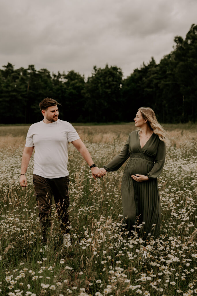 Werdende Eltern spazieren Hand in Hand durch eine Blumenwiese während ihres Schwangerschaftsshootings.