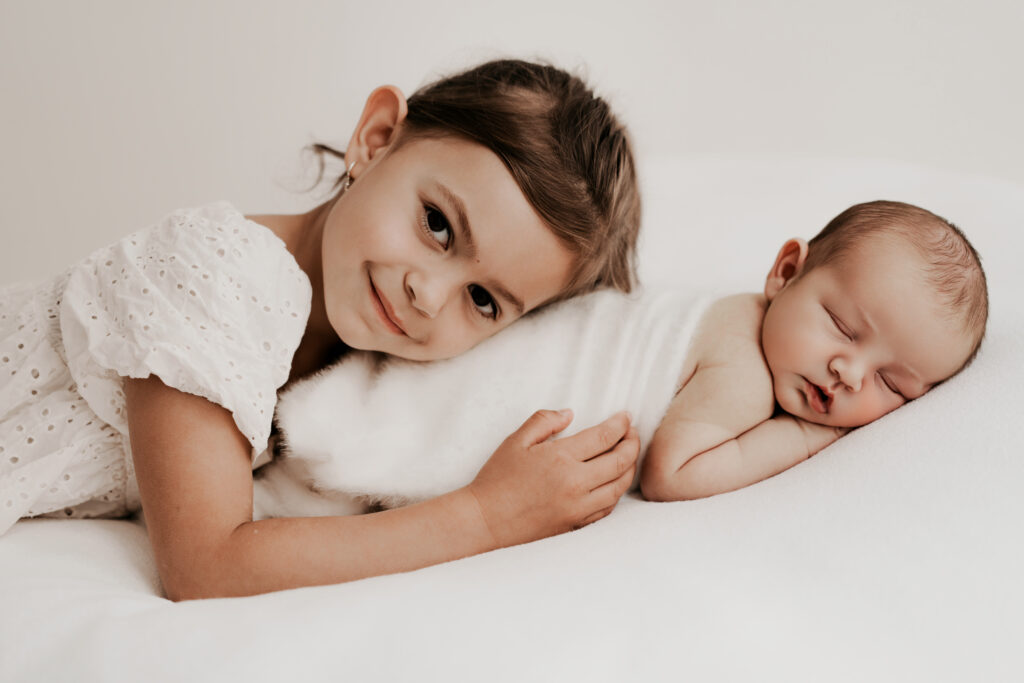 Natürliches Geschwisterfoto mit Baby und großer Schwester auf einem hellen weißen Bett.