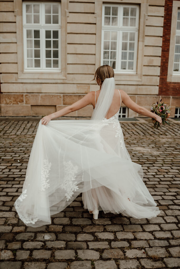 Braut dreht sich mit ihrem Hochzeitskleid in der Hand vor dem Schloss Münster